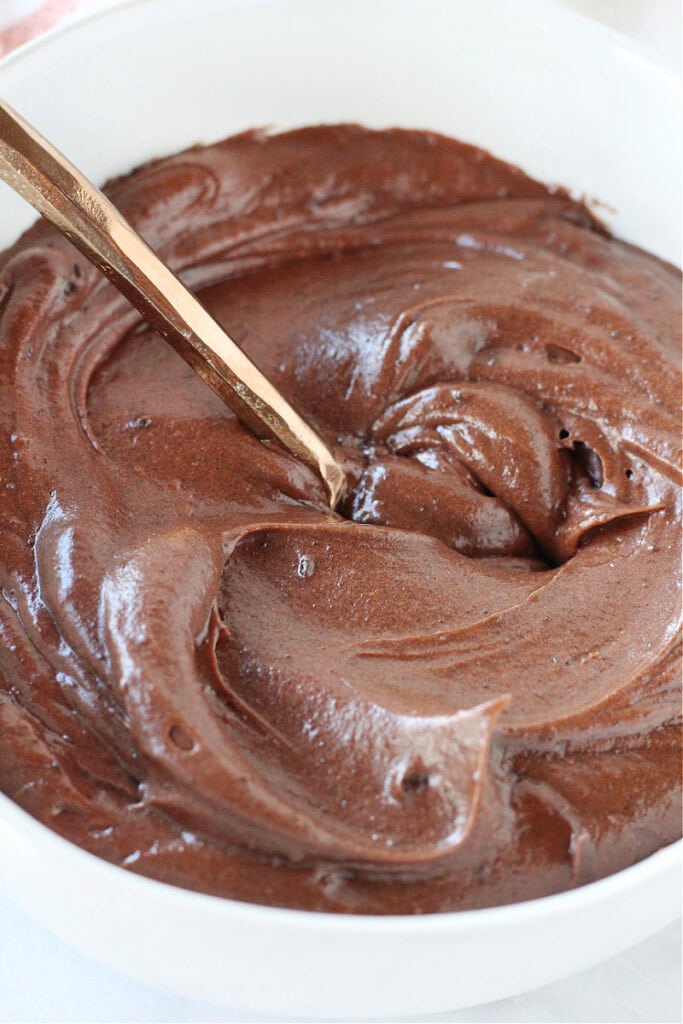Chocolate frosting in a white bowl with a copper spoon.