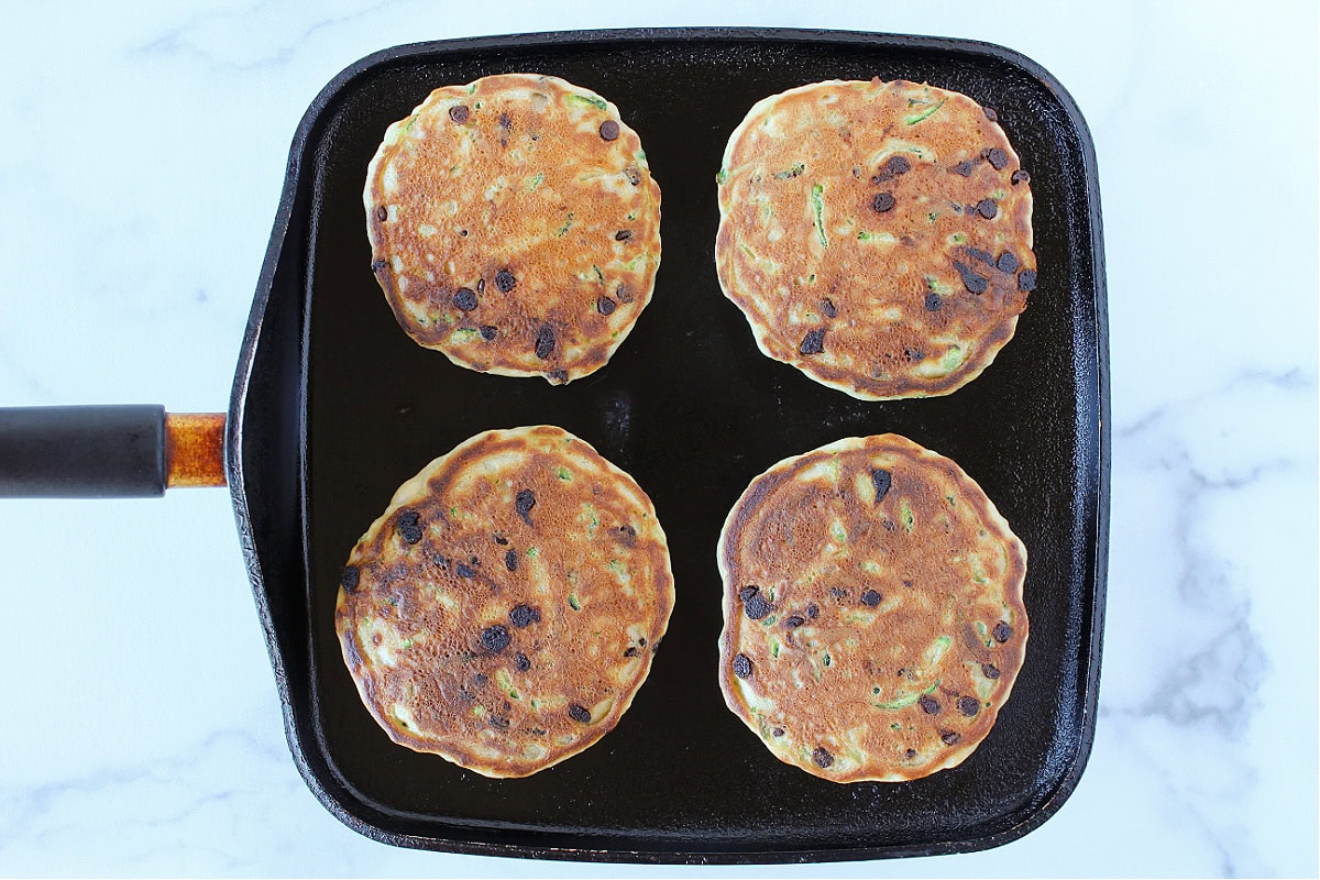 Four zucchini pancakes with mini chocolate chips on a black griddle.