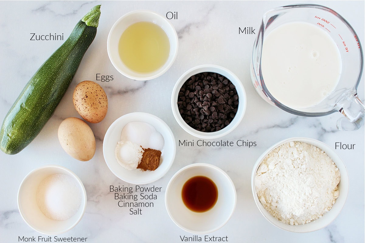 Labeled ingredients for chocolate chip zucchini pancakes on a white marble countertop.