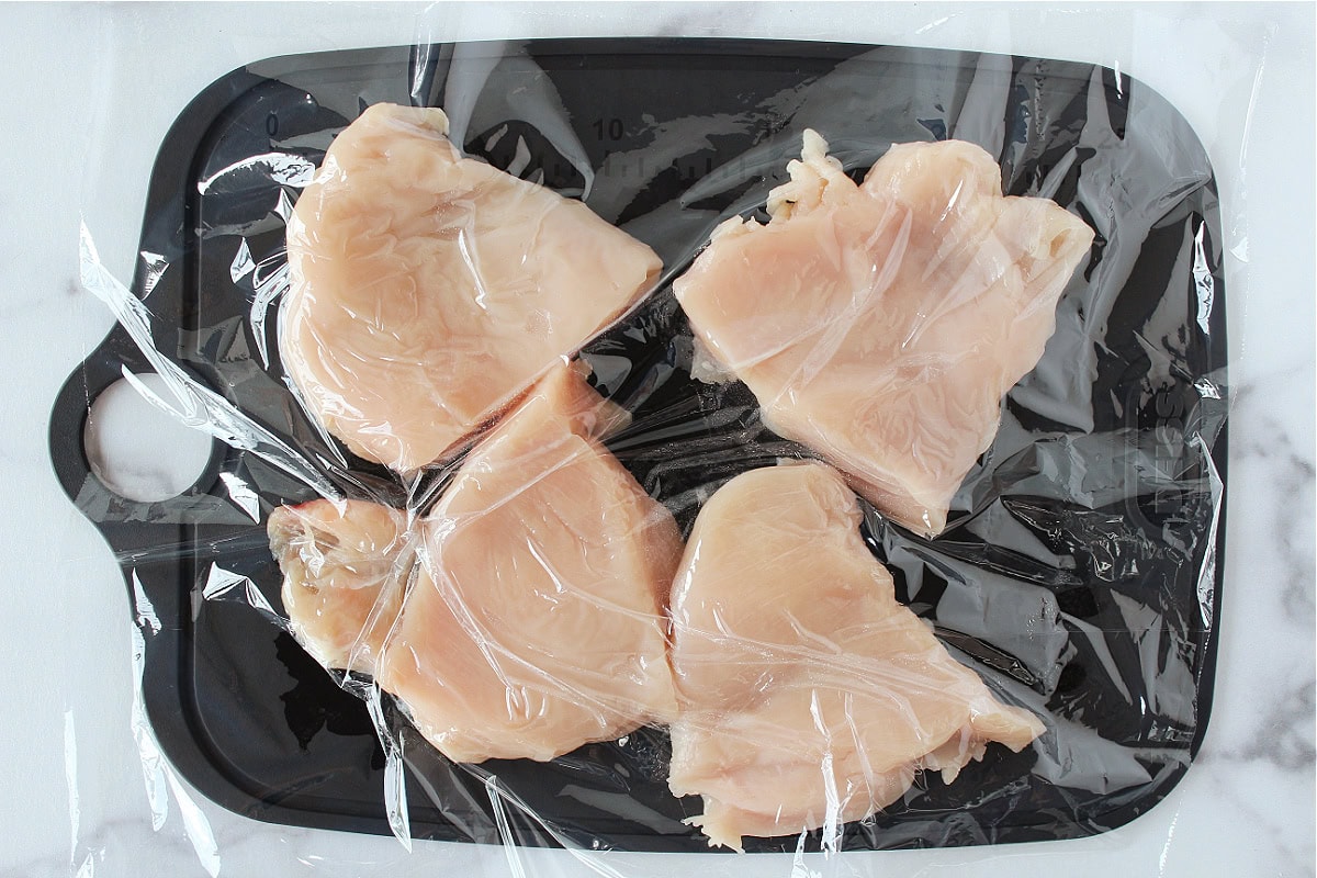 Four chicken breasts on a cutting board topped with plastic wrap.