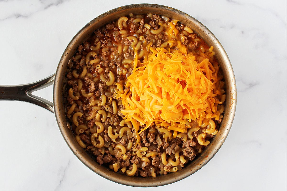 Shredded cheese on top of cooked elbow noodles and ground beef in a pan.