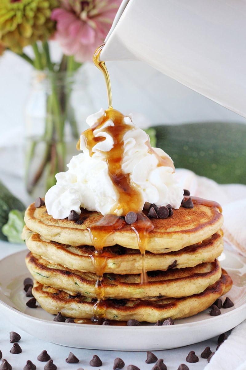 Maple syrup pouring over pancakes topped with whipped cream and mini chocolate chips.