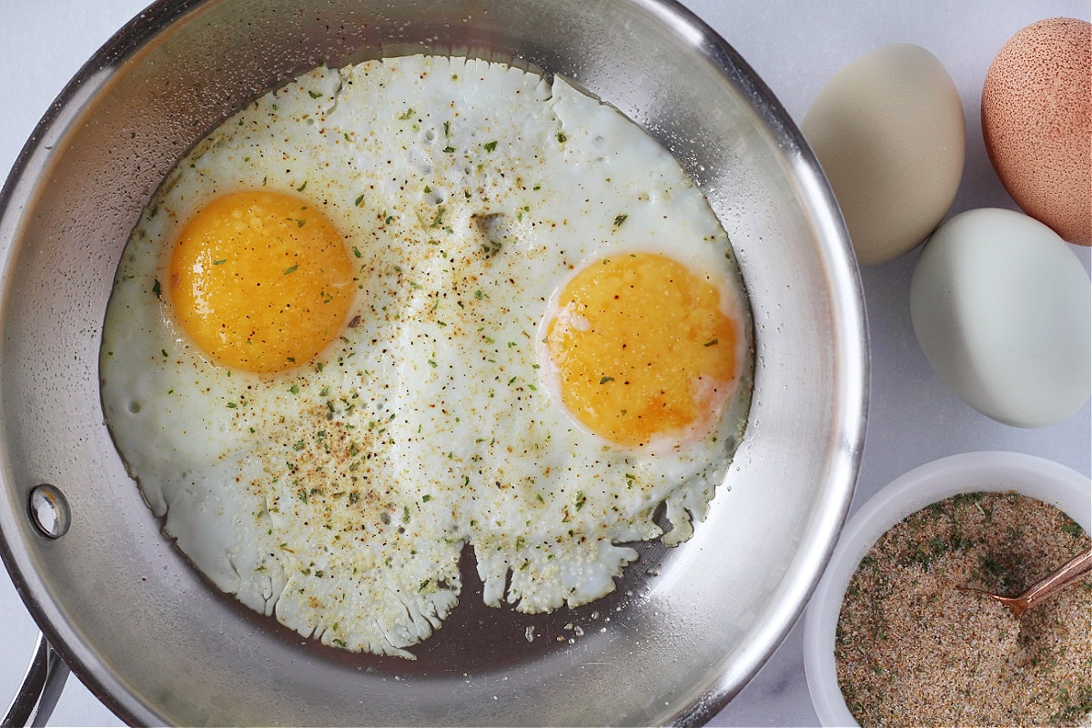 Two eggs cooked over easy in a pan next to three eggs and burger seasoning.