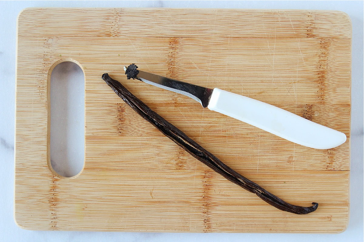 A knife with vanilla bean paste on a cutting board next to a vanilla bean.
