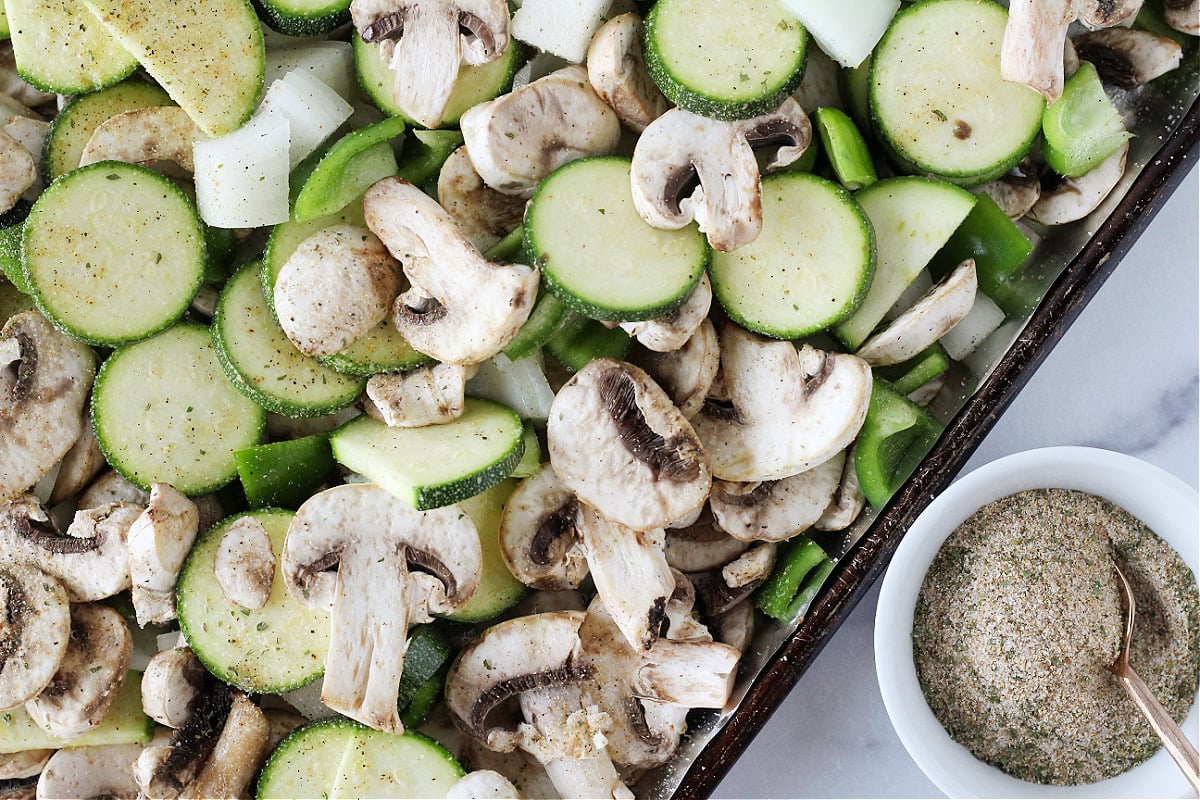Sliced zucchini, mushrooms, onion and bell pepper on a sheet pan with seasoning.