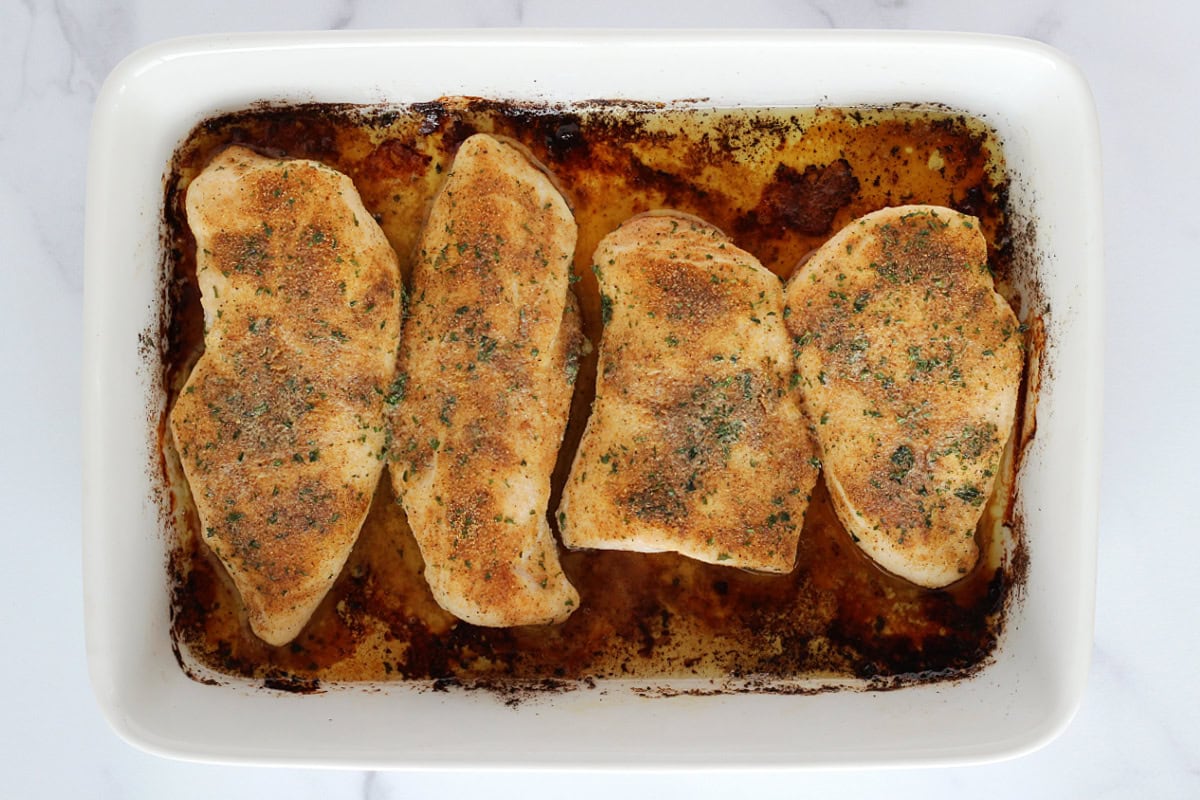 Four baked chicken breasts with seasoning in a white baking dish.
