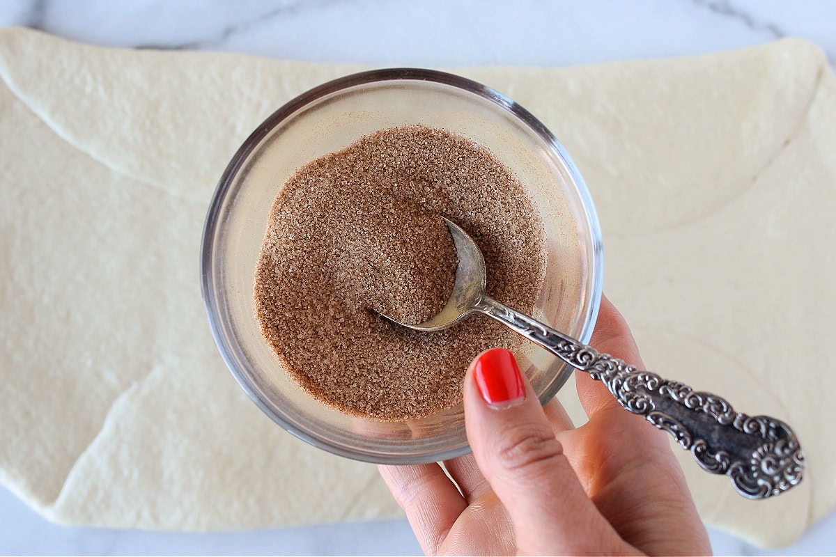 Cinnamon sugar in a bowl with a spoon over bread dough rolled flat.