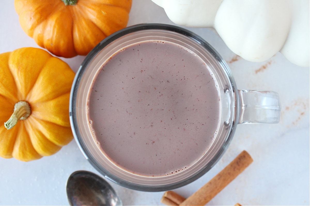 Pumpkin spice hot chocolate in a glass mug surrounded by pumpkins and cinnamon sticks.