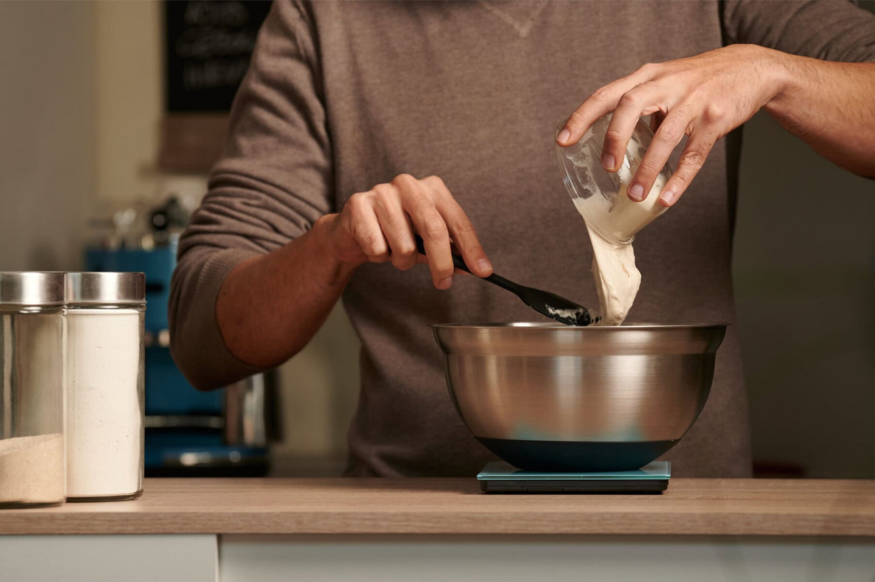 A man pouring sourdough starter into a metal bowl on a kitchen scale.