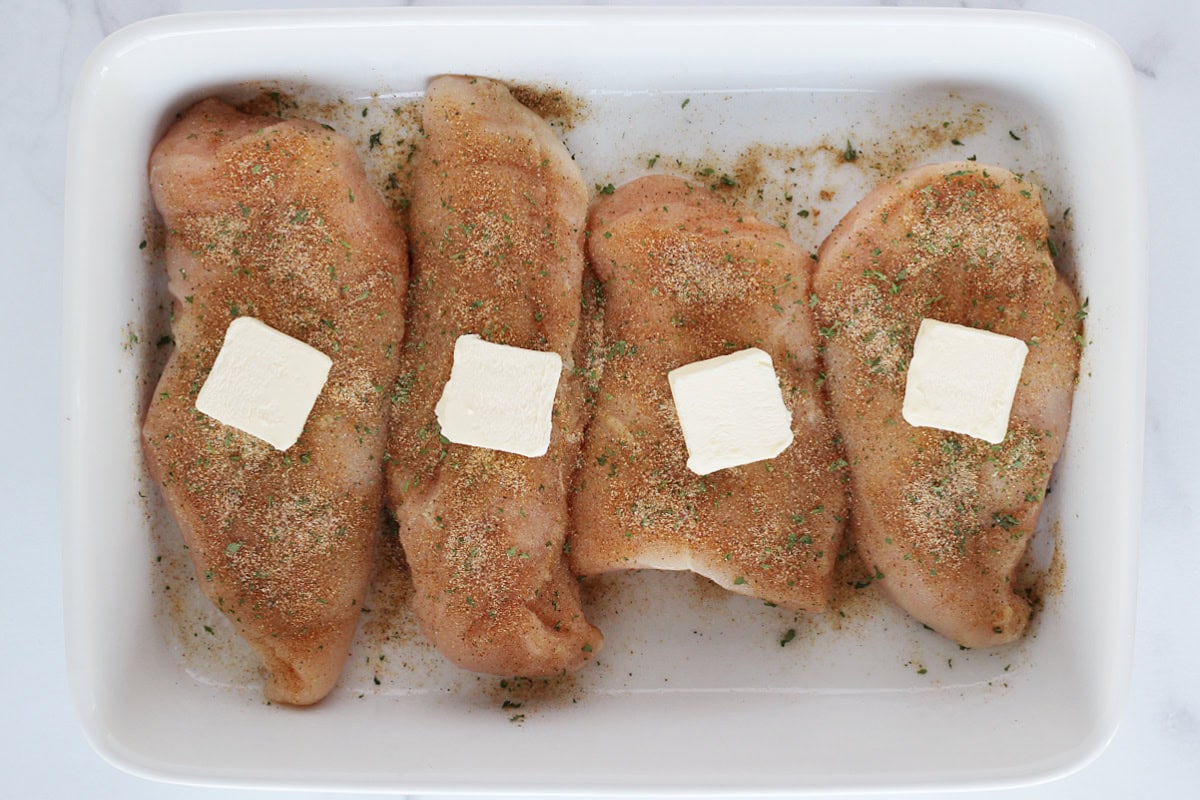 Four raw chicken breasts seasoned and topped with a tab of butter in the pan.