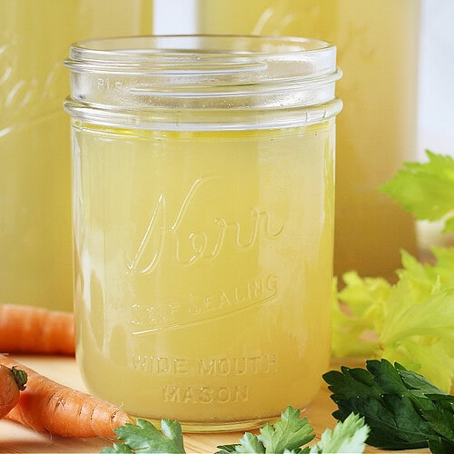 Mason jars filled with chicken broth surrounded by carrots and celery.
