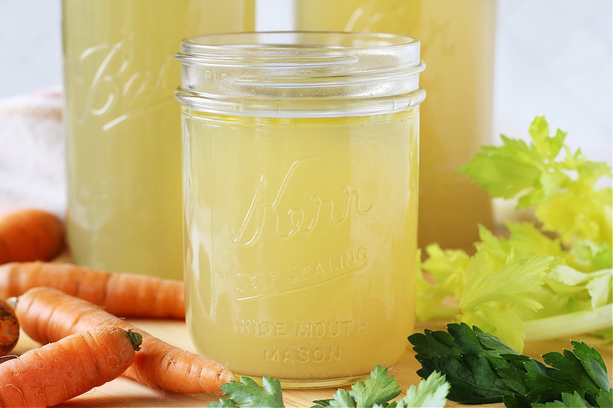 Mason jars filled with chicken broth surrounded by carrots and celery.