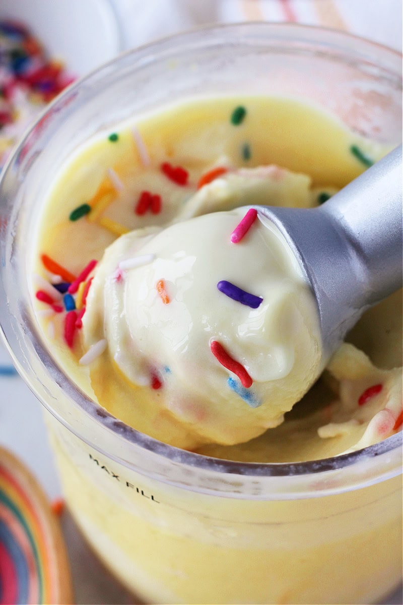 Metal ice cream scoop with rainbow sprinkles on top in clear container.
