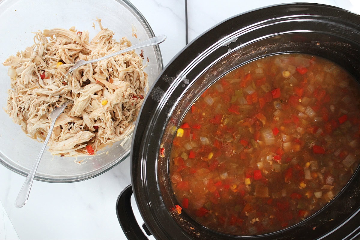 Shredded chicken in a glass bowl with forks beside a slow cooker with soup.