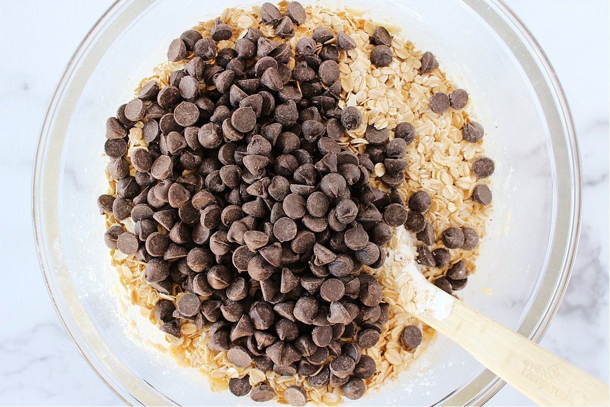 Chocolate chips and rolled oats on cookie batter in a glass mixing bowl.