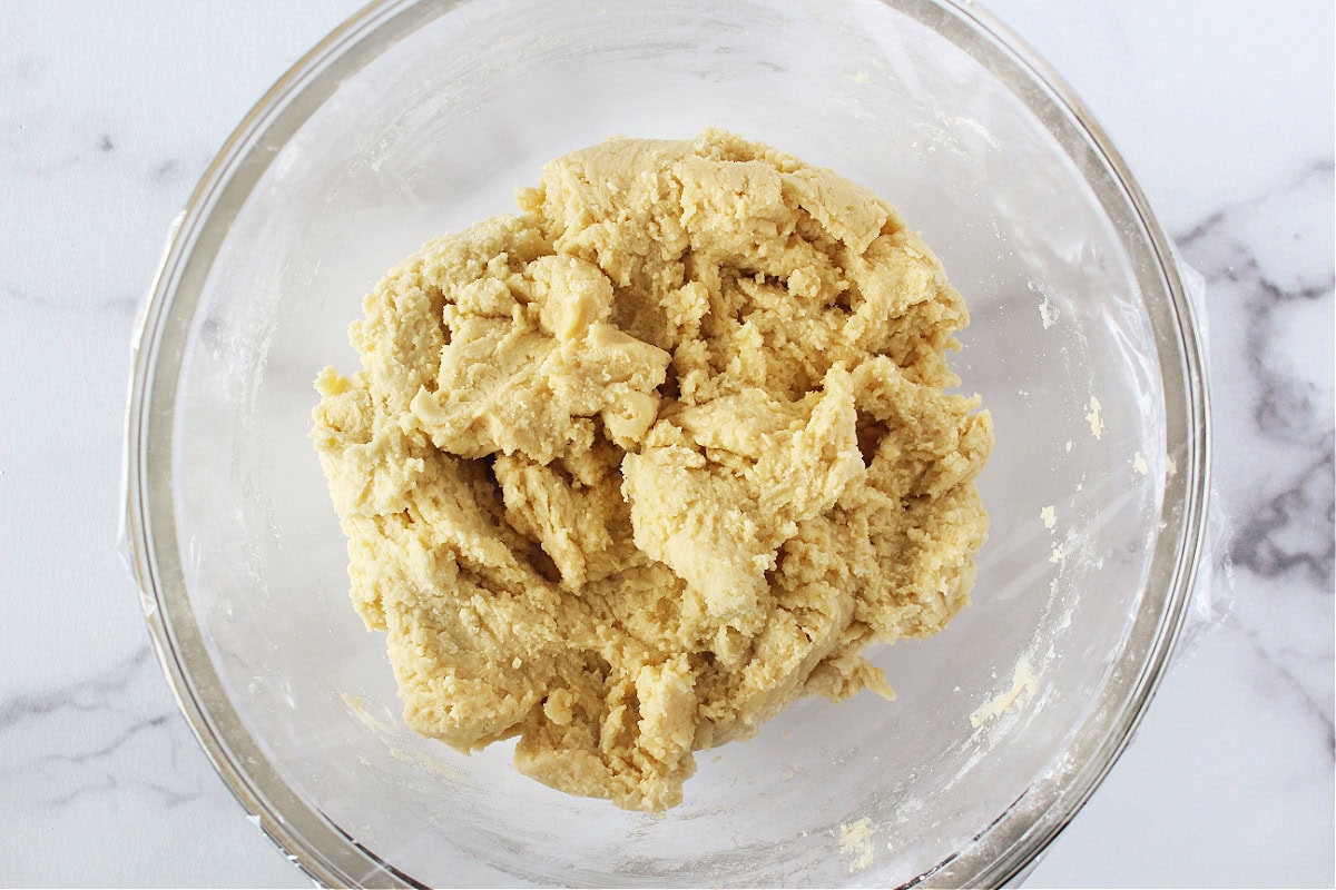 Sugar cookie dough in a glass mixing bowl covered with plastic wrap.