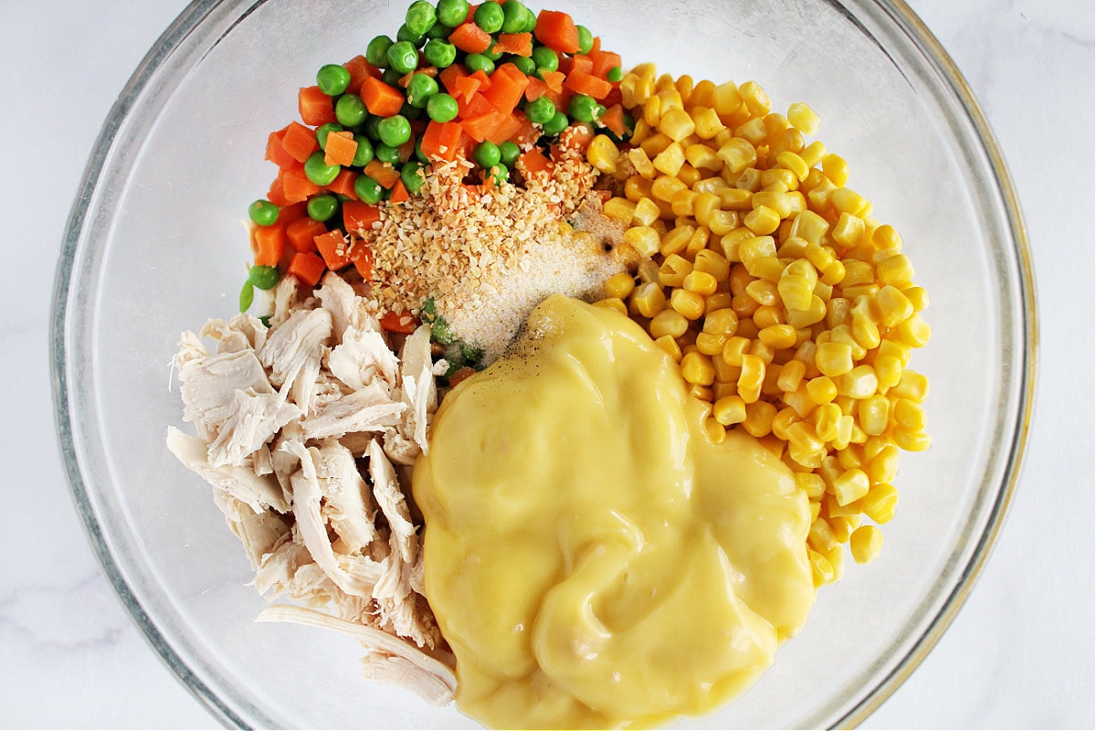 Peas, carrots, corn, chicken, cream of mushroom soup, and seasonings in a glass bowl.