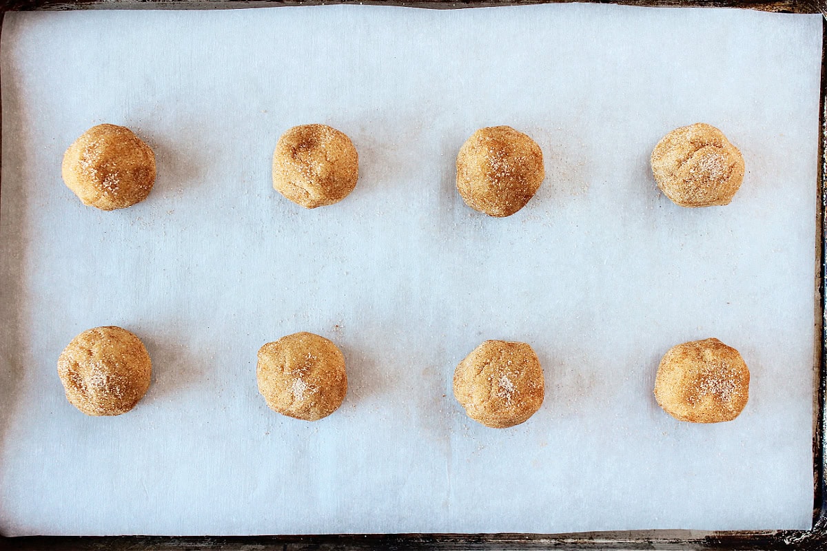 Eight cookie dough balls coated in cinnamon sugar on a lined baking sheet.