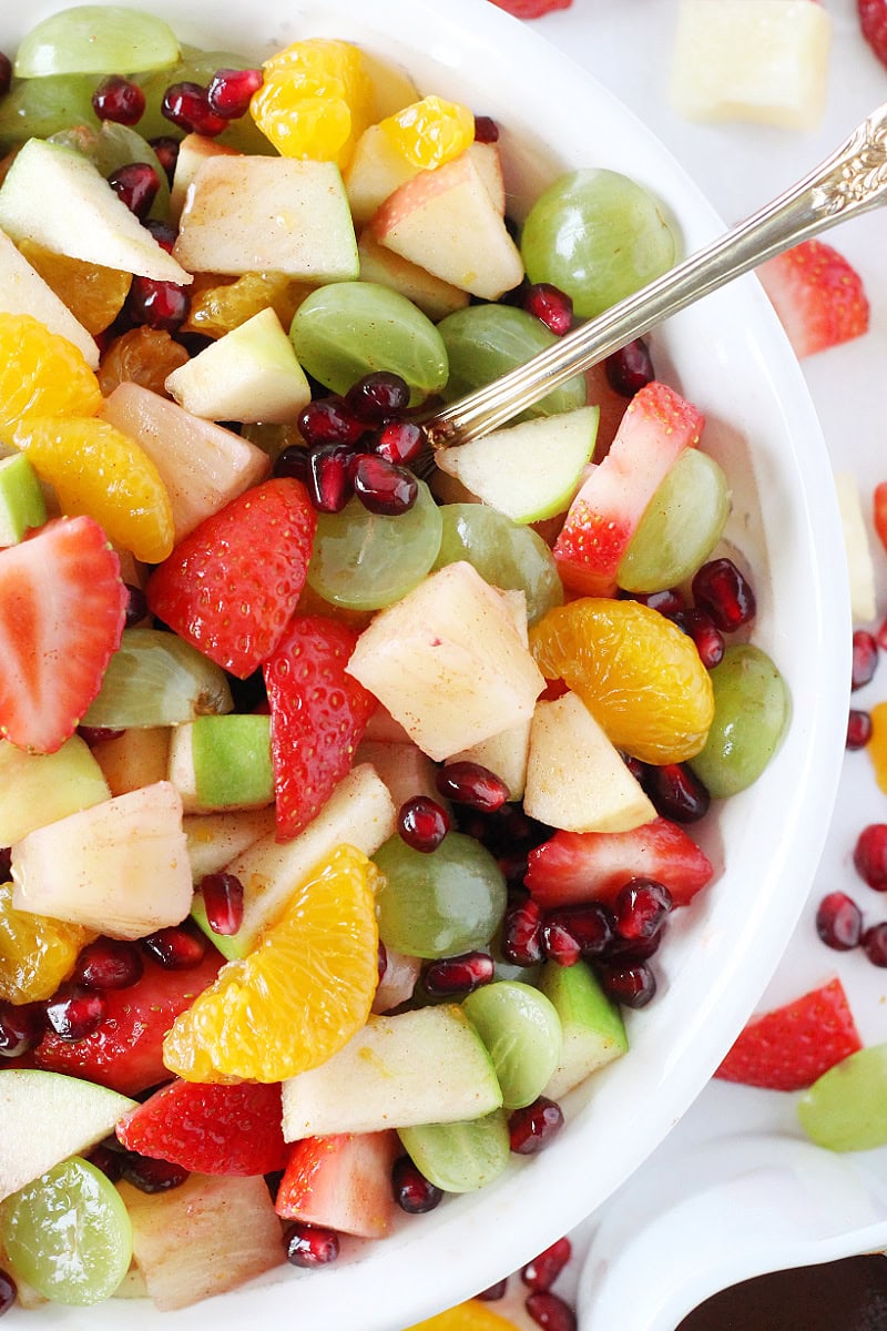 A gold spoon in a bowl of winter fruit salad with pineapple, apples, and mandarin oranges.