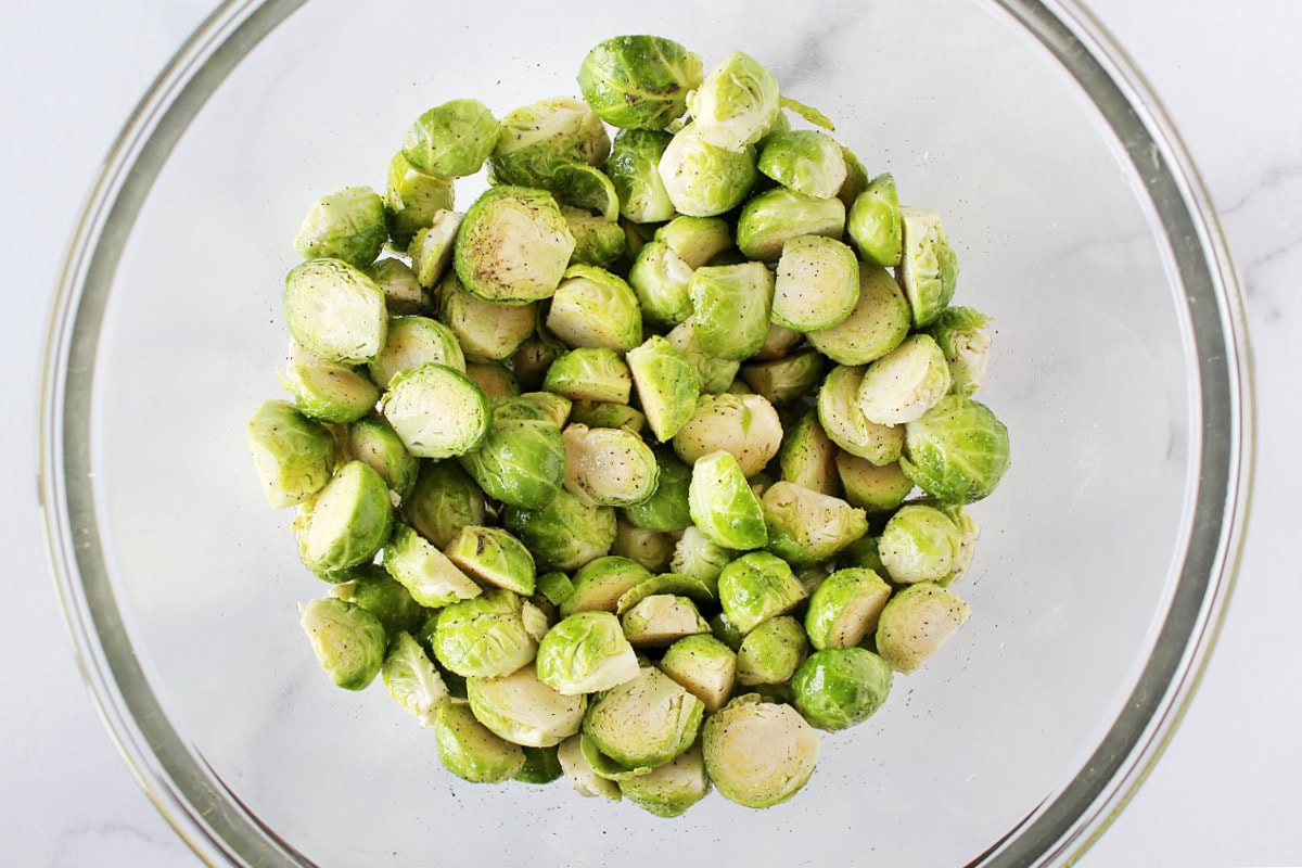 Halved Brussel sprouts in a glass bowl coated with salt and black pepper.