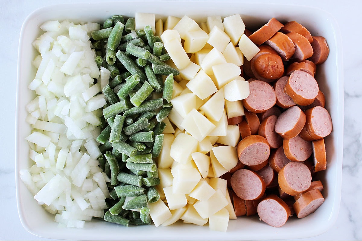 Diced onion, frozen green beans, diced potatoes, and sliced sausage in a white baking dish.