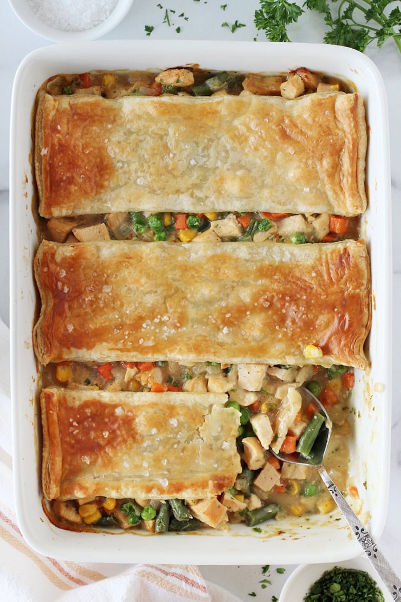 A spoon in a 9x13 white pan of homemade chicken pot pie with puff pastry.