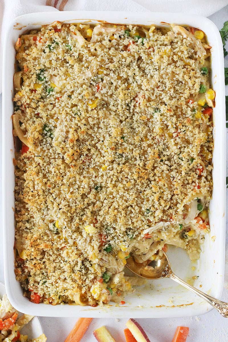 A portion of chicken noodle casserole missing from the white baking dish with gold spoon.