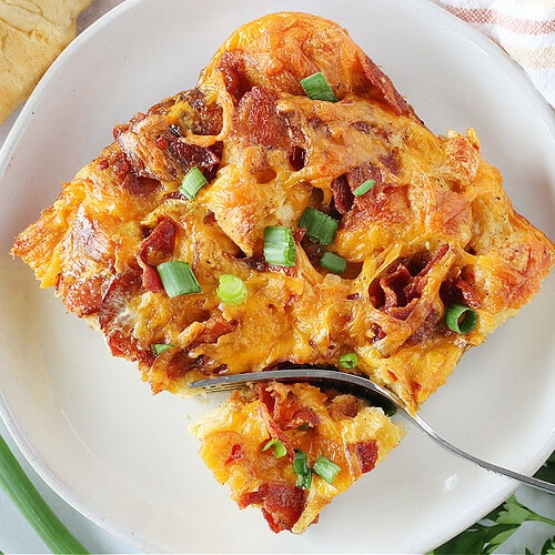 A fork slicing into a portion of croissant breakfast casserole on a white plate.
