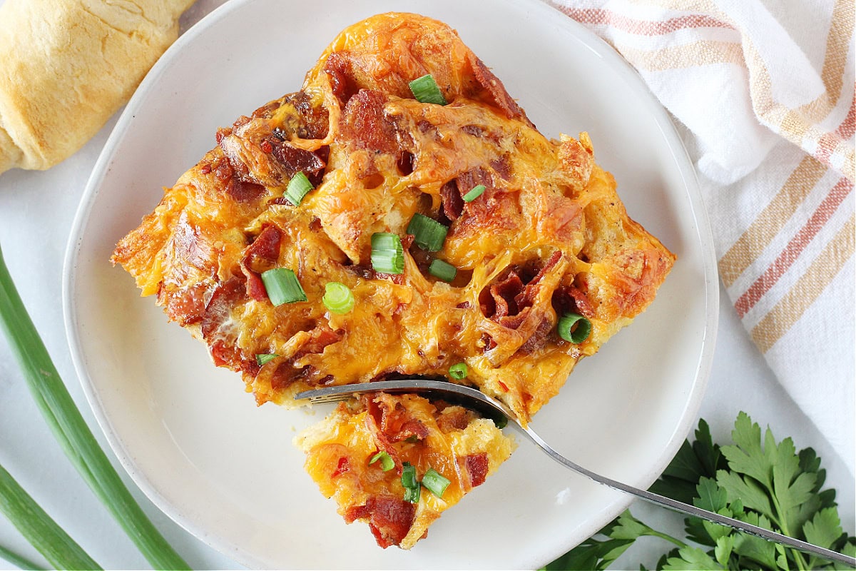 A fork slicing into a portion of croissant breakfast casserole on a white plate.