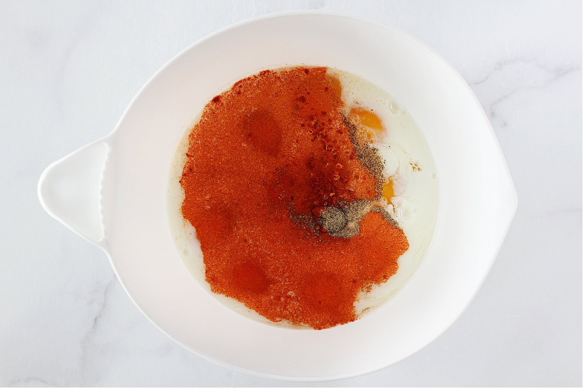 Paprika and black pepper on top of eggs and milk in a mixing bowl.