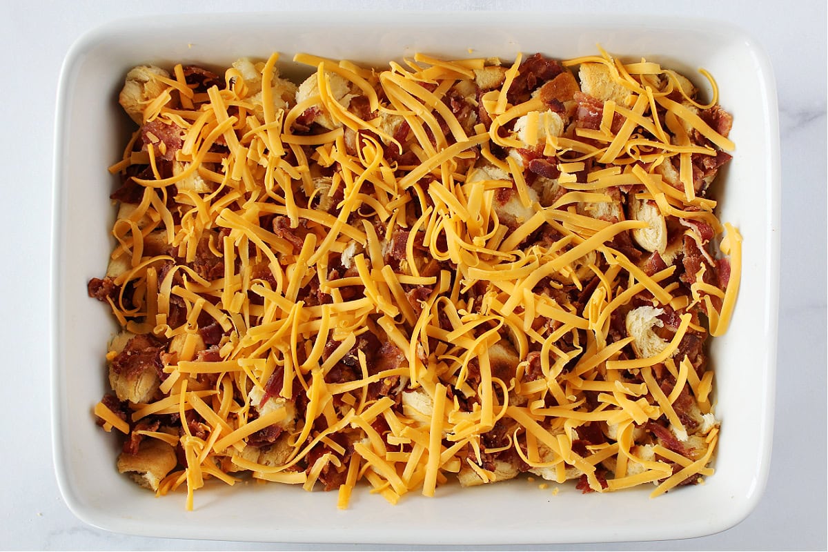 Shredded cheese and bacon on top of croissant pieces in a baking dish.