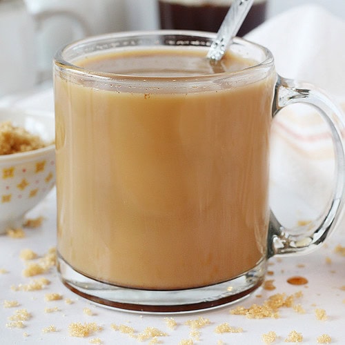 A latte in a glass mug with a spoon surrounded by brown sugar.