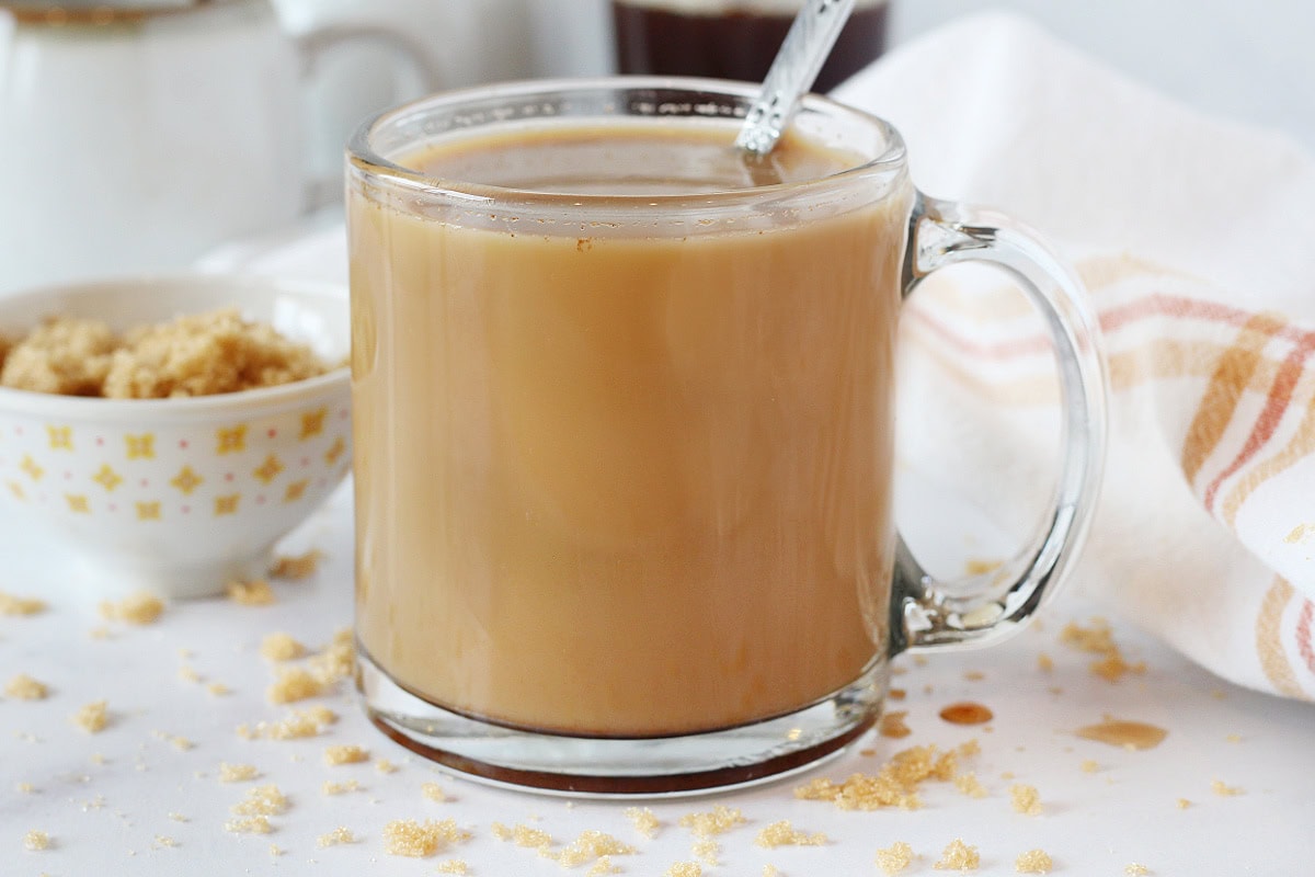 A latte in a glass mug with a spoon surrounded by brown sugar.