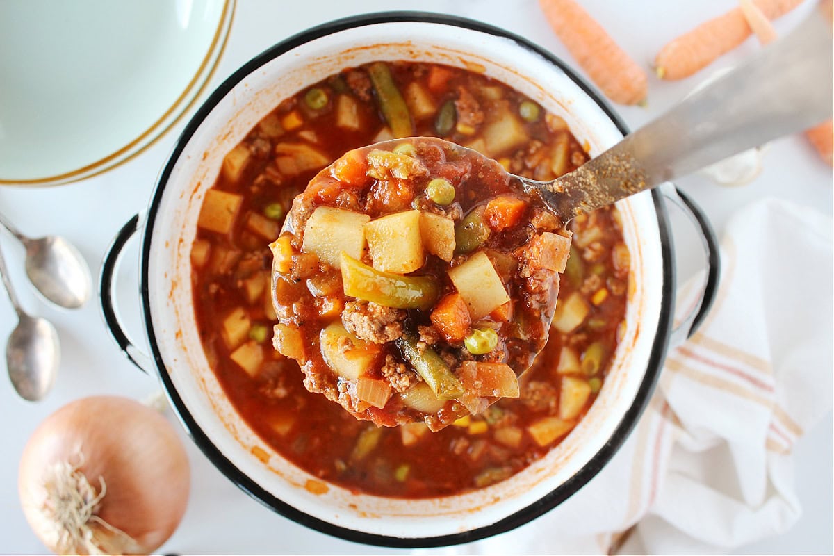 A metal ladle full of hamburger soup over the soup pot.