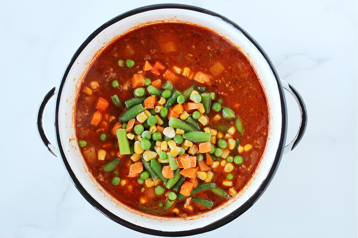 Frozen mixed vegetables on top of hamburger soup in a white pot.