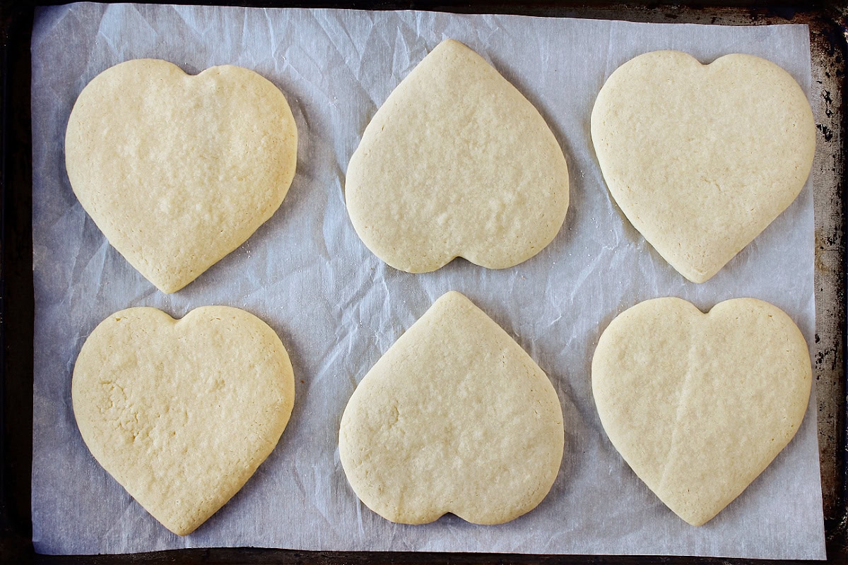 Six baked sugar cookies shaped liked hearts on a parchment paper lined baking sheet.