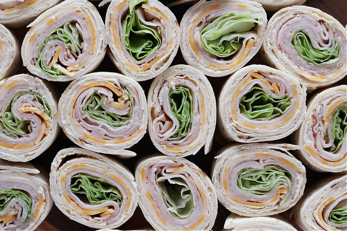 Roll ups with turkey lunch meat, cheese slices, and lettuce on a cutting board.