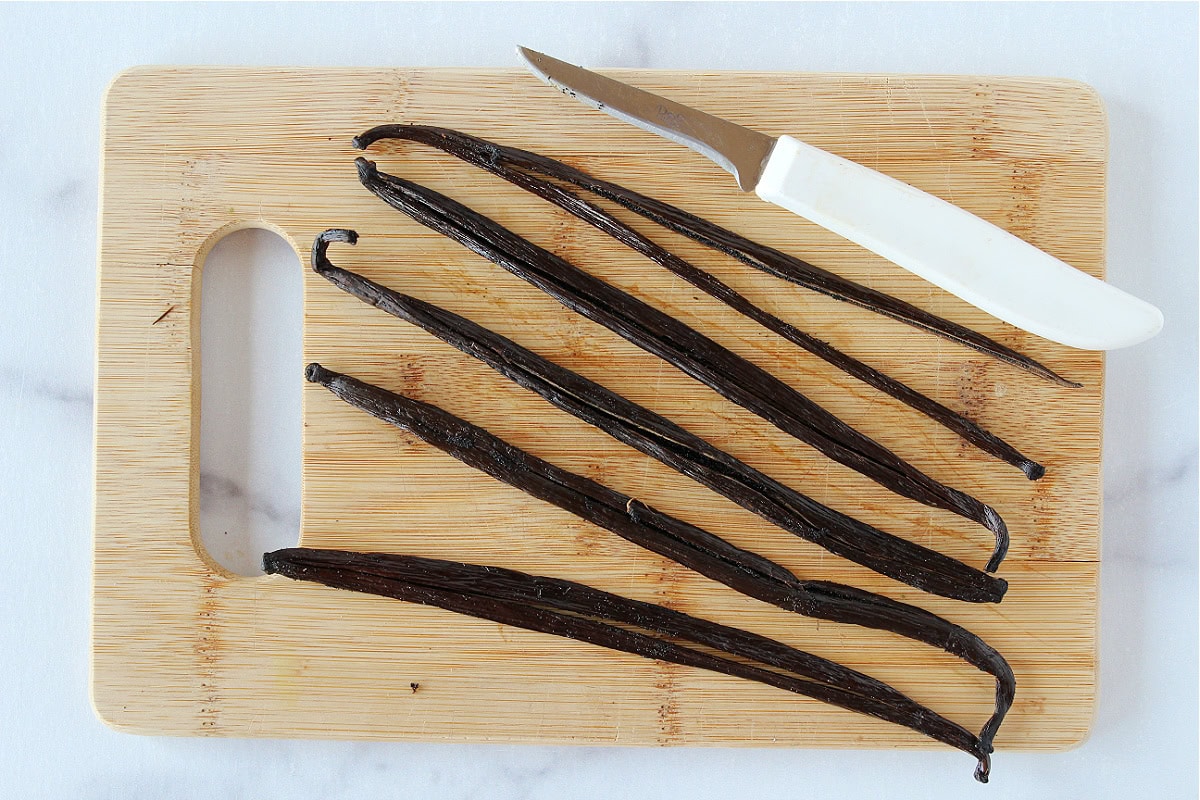 Whole vanilla beans on a wooden cutting board with a pairing knife.