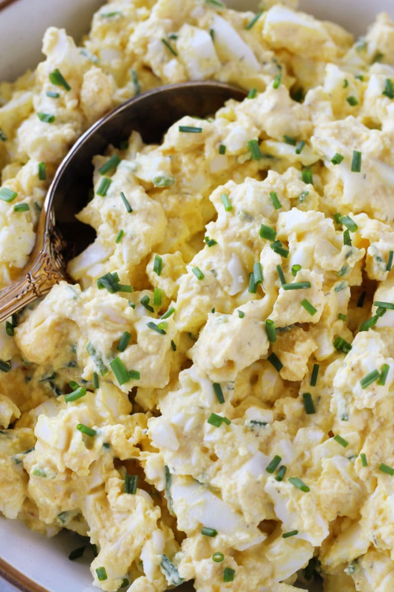 A golden spoon in a bowl of creamy egg salad topped with chives.