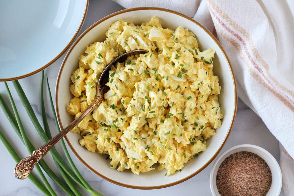 Egg salad topped with chopped chives in a white bowl with gold spoon.