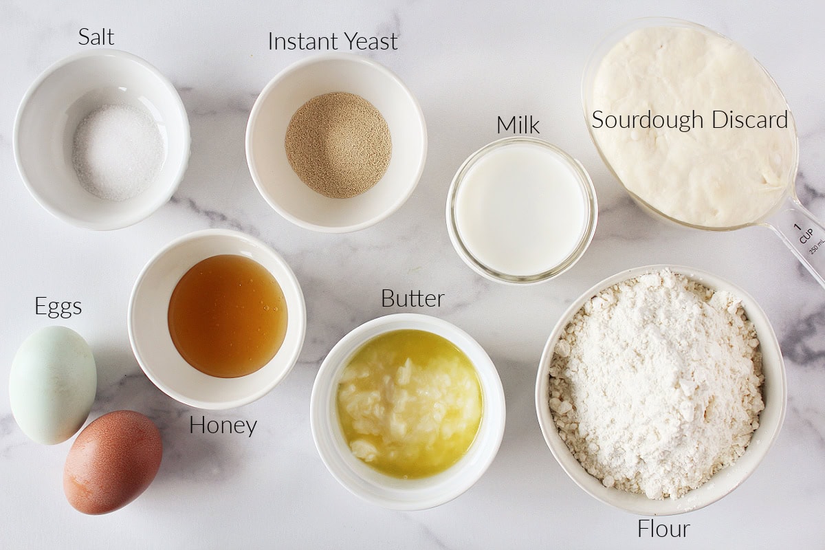 Labeled ingredients for soft and fluffy sourdough rolls on a white marble countertop.