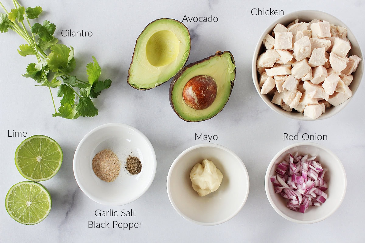 Labeled ingredients for creamy chicken avocado chicken salad on white marble countertop.