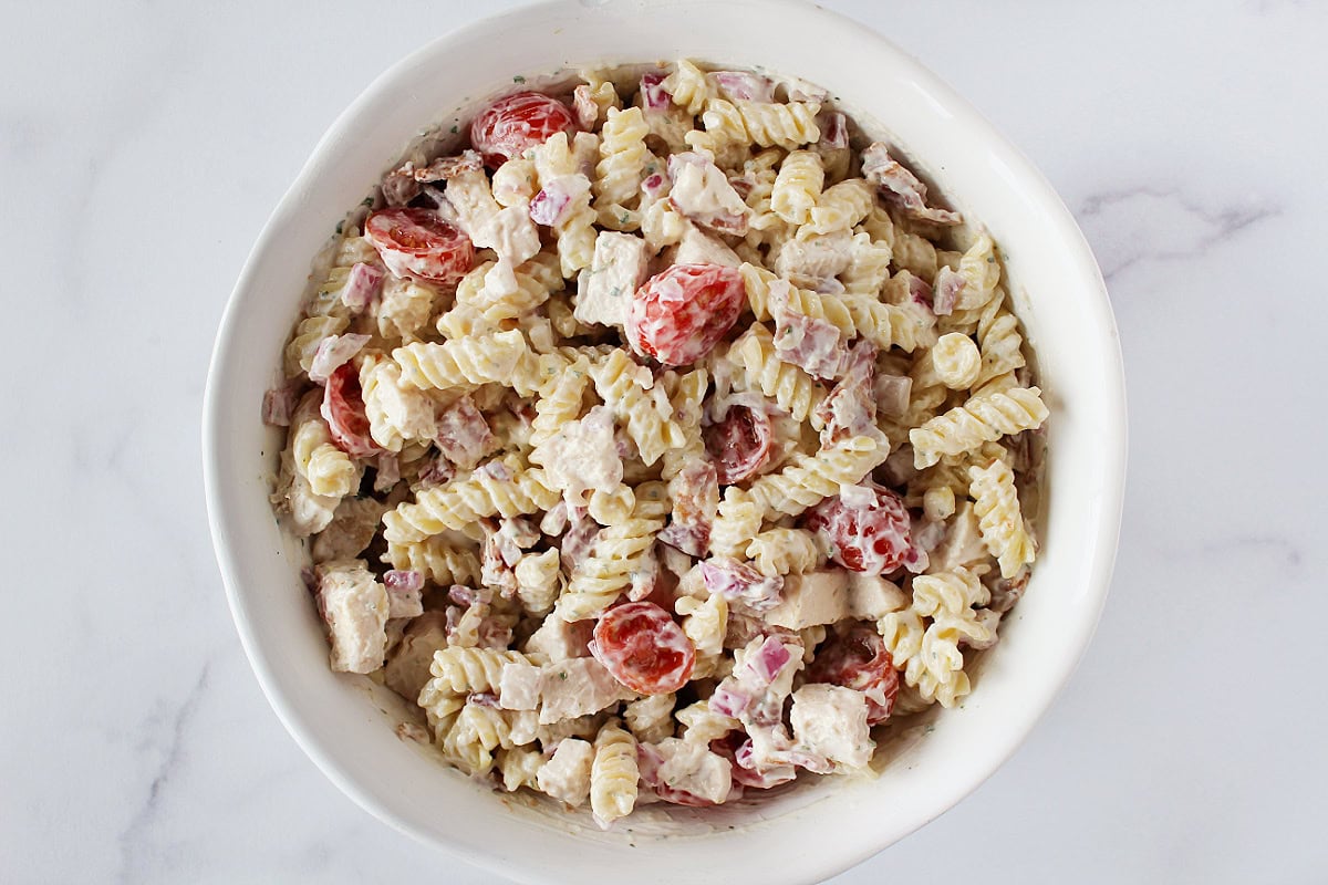 Rotini pasta, halved grape tomatoes, bacon and plain Greek yogurt mixed in a white bowl.