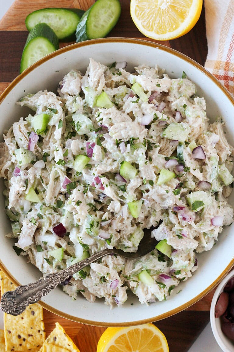 Tzatziki chicken salad in a bowl on a cutting board with cucumbers, lemon, and tortilla chips.