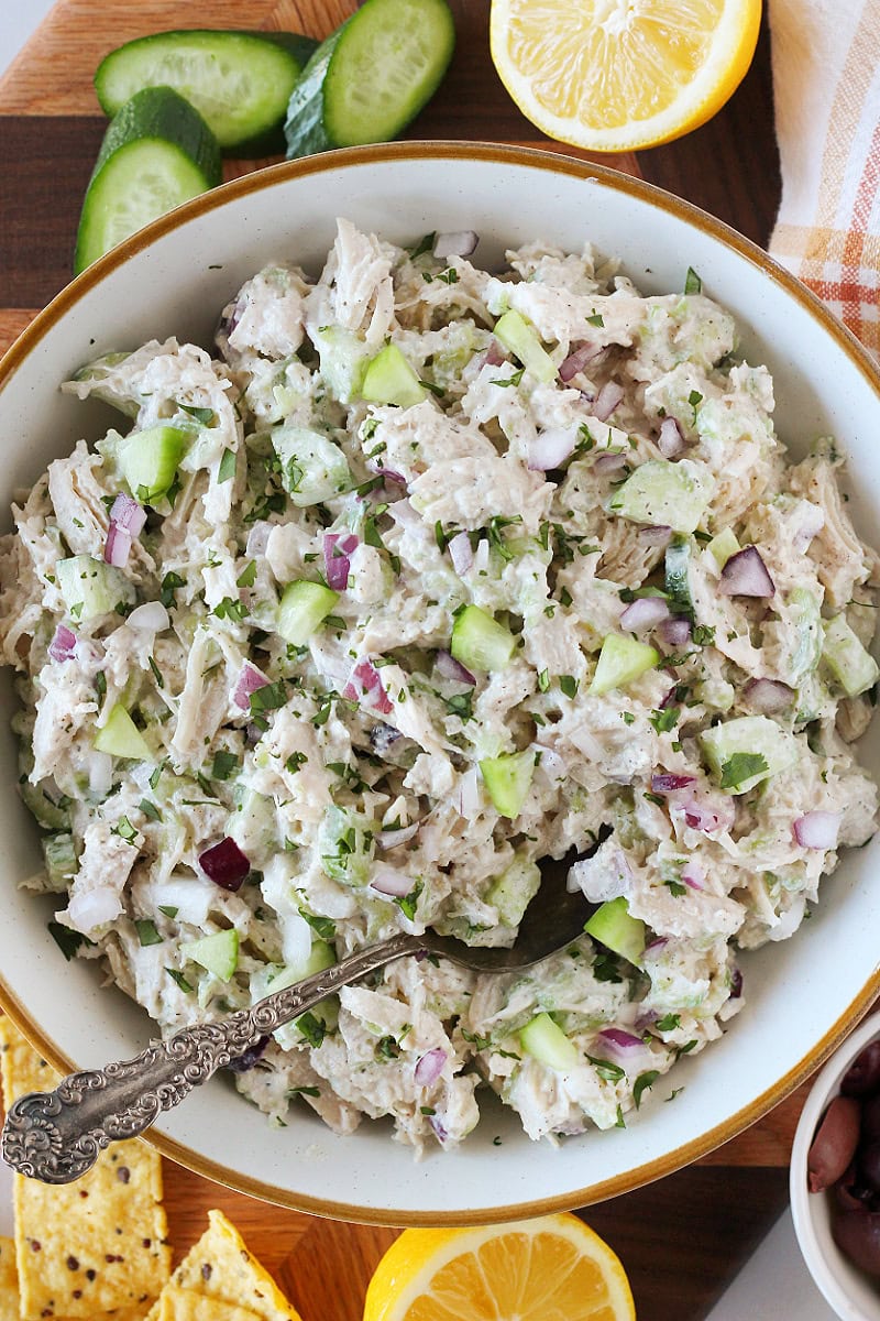 Tzatziki chicken salad in a bowl on a cutting board with cucumbers, lemon, and tortilla chips.