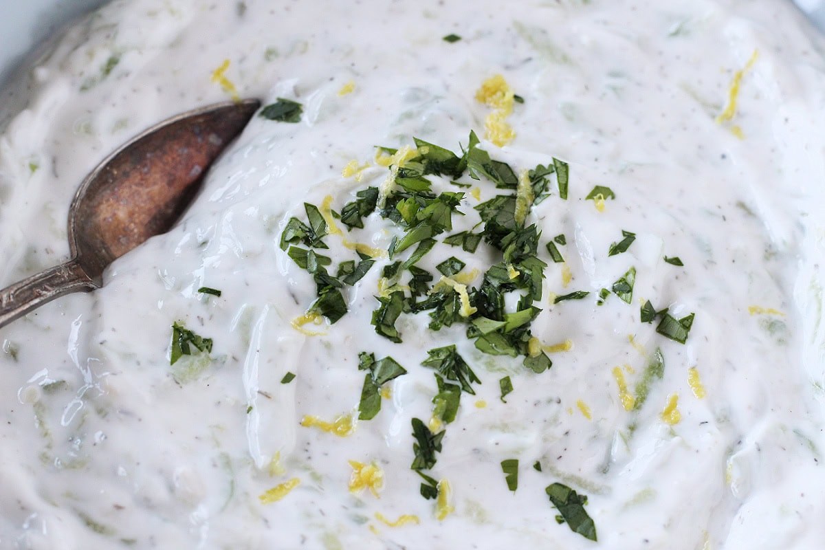 A spoon in a bowl of tzatziki topped with lemon zest and parsley.