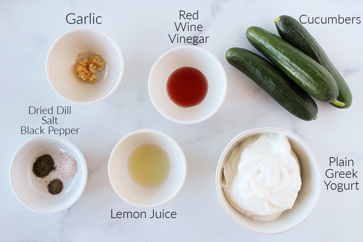 Labeled ingredients for homemade tzatziki on a white marble countertop.