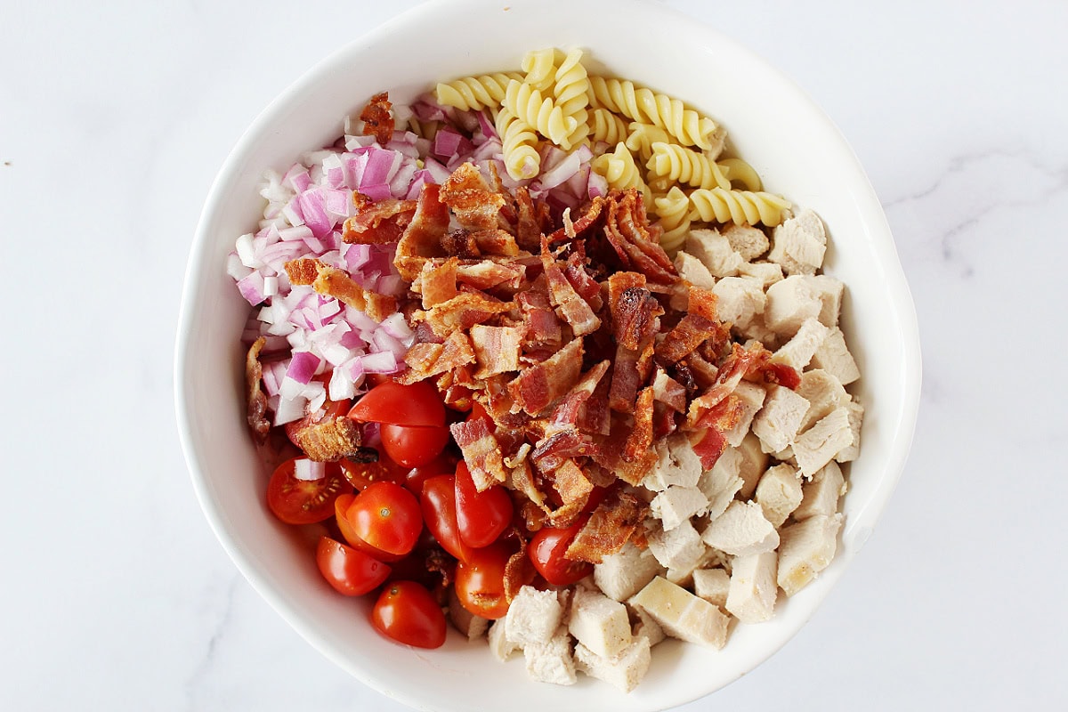 Cooked rotini pasta, halved grape tomatoes, diced chicken breast and bacon in bowl.