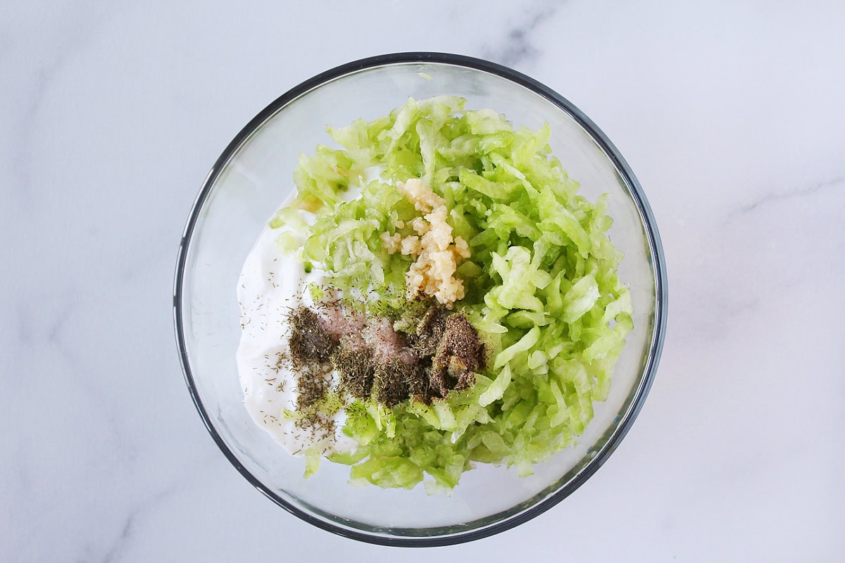 Shredded cucumber, minced garlic, plain Greek yogurt, and seasonings in a glass bowl.