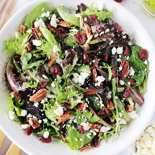 Mixed greens in a bowl topped with crumbled feta cheese, dried cranberries, and pecans.